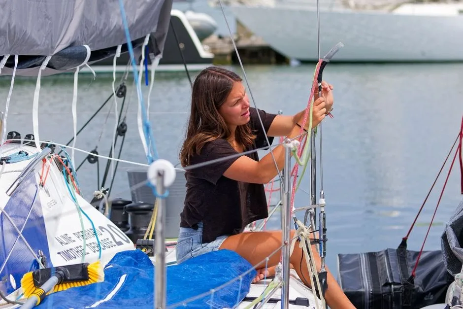Le voilier SV Liahona navigue au large, voiles déployées, sous un ciel gris avec un membre d'équipage à bord